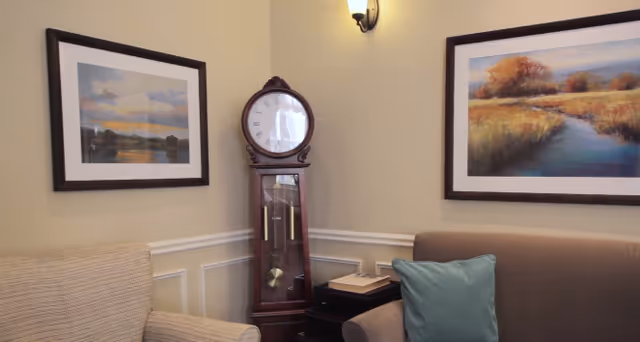 A cozy corner of a living room featuring a beige armchair, a brown sofa with a teal pillow, a wooden grandfather clock, two framed landscape paintings on the walls, and a wall-mounted light fixture above.