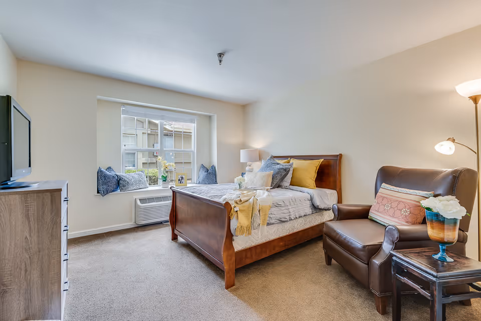 Bright furnished bedroom with a wooden sleigh bed, armchair, TV on a dresser, and a large window seat.