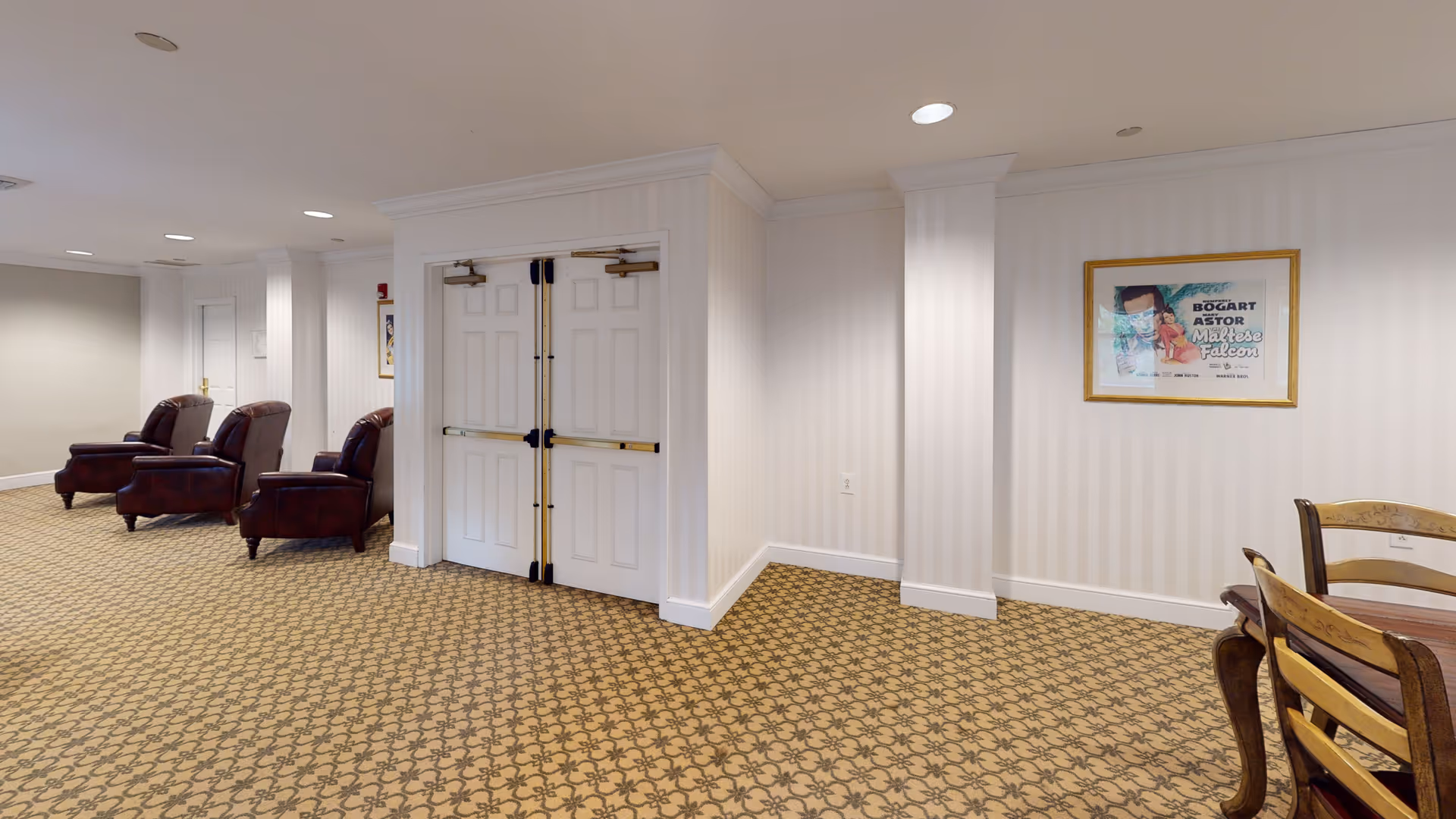 Interior view of a senior living facility common area with patterned carpet, three brown leather armchairs along the left wall, a double white door in the center, and a framed vintage movie poster on the right wall above a wooden dining table with chairs.