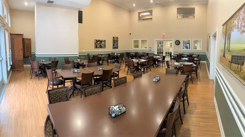 A spacious dining room with multiple wooden tables and chairs arranged neatly. The room has light-colored walls with green wainscoting, wooden flooring, and large windows letting in natural light. There are decorative paintings on the walls and a door at the far end of the room.