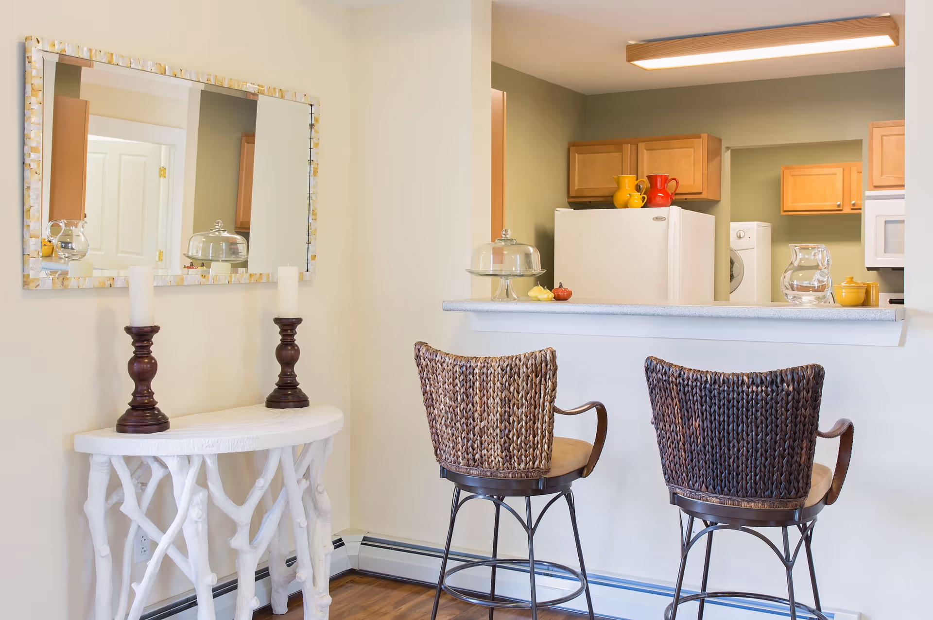 Interior view of a small kitchen and dining area with two woven bar stools at a counter. The kitchen has wooden cabinets, a white refrigerator, and a washing machine visible in the background. A white console table with two dark candle holders and a large mirror above it is positioned against the wall.