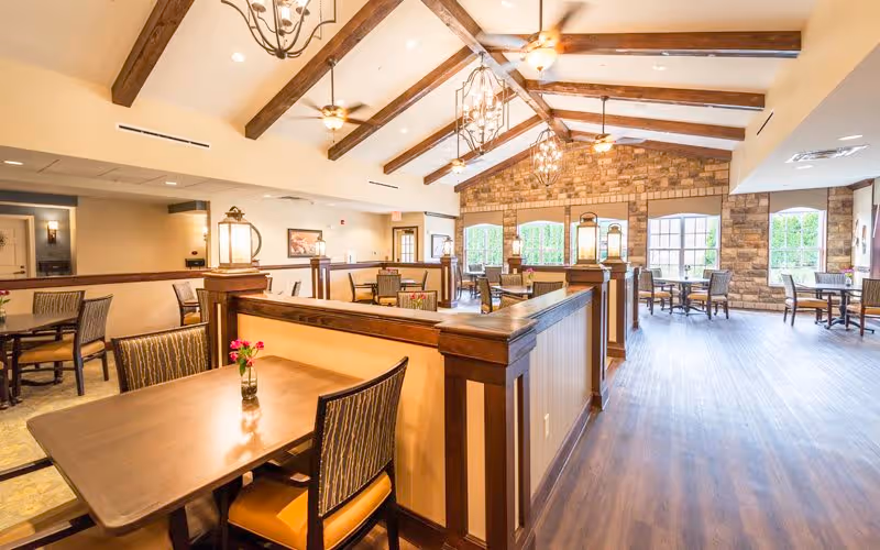 Bright and spacious dining area with wooden tables and chairs, decorative ceiling beams, large windows, and stone accent walls. The room features warm lighting fixtures and a vase with flowers on the closest table.