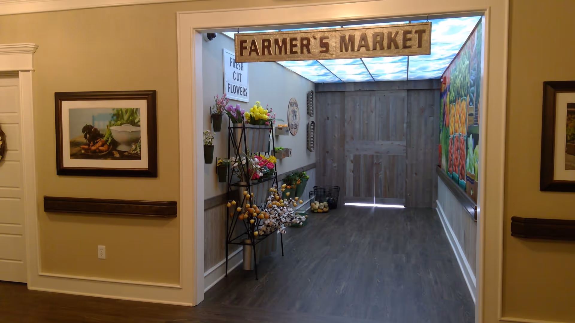Indoor hallway area decorated as a farmer's market with a hanging sign that reads 'FARMER'S MARKET'. There are shelves with artificial flowers and plants on the left side, a sign that says 'FRESH CUT FLOWERS', and a mural of various vegetables on the right wall. The floor is dark wood, and the ceiling has a sky-themed light panel. The walls are beige with framed pictures on either side of the entrance.