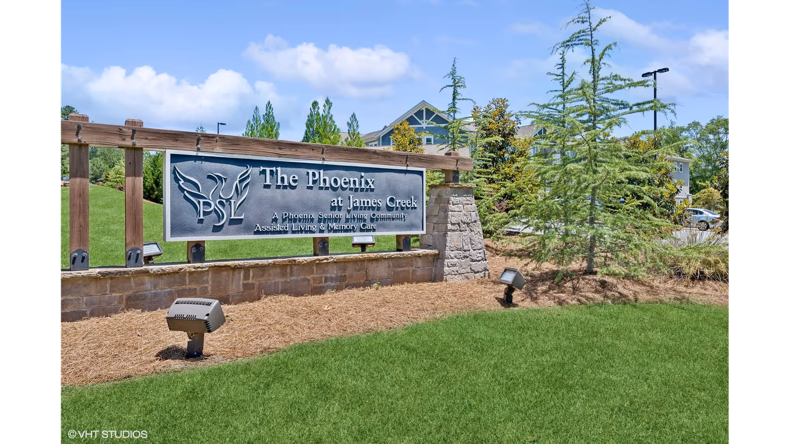 Outdoor view of the entrance sign for The Phoenix at James Creek, a senior living community offering assisted living and memory care. The sign is mounted on a stone and wood structure with landscaping including green grass, pine trees, and a clear blue sky in the background.