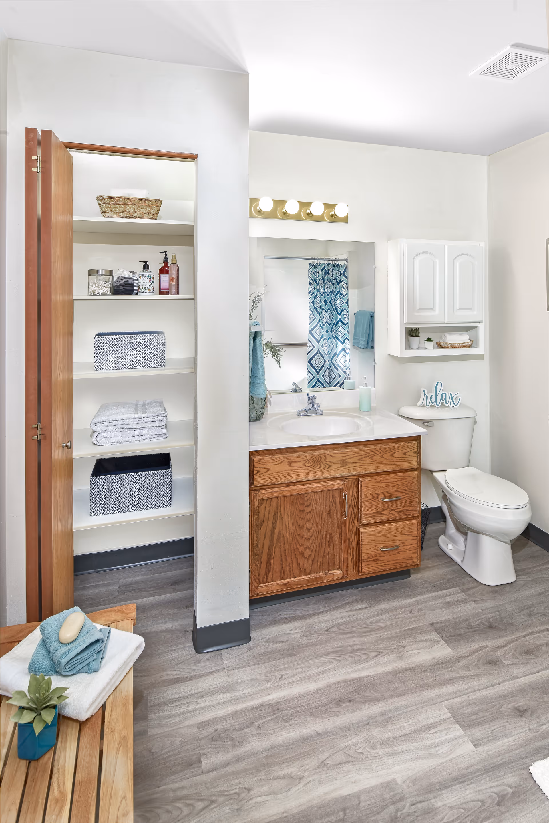 A clean and well-organized bathroom featuring a wooden vanity with a sink, a large mirror with lights above it, a white toilet with a decorative 'relax' sign on the tank, and a white wall cabinet above the toilet. To the left, there is an open closet with shelves holding towels, baskets, and toiletries. The floor is covered with light gray wood-style flooring, and a wooden bench with folded towels and a small plant is visible in the foreground.