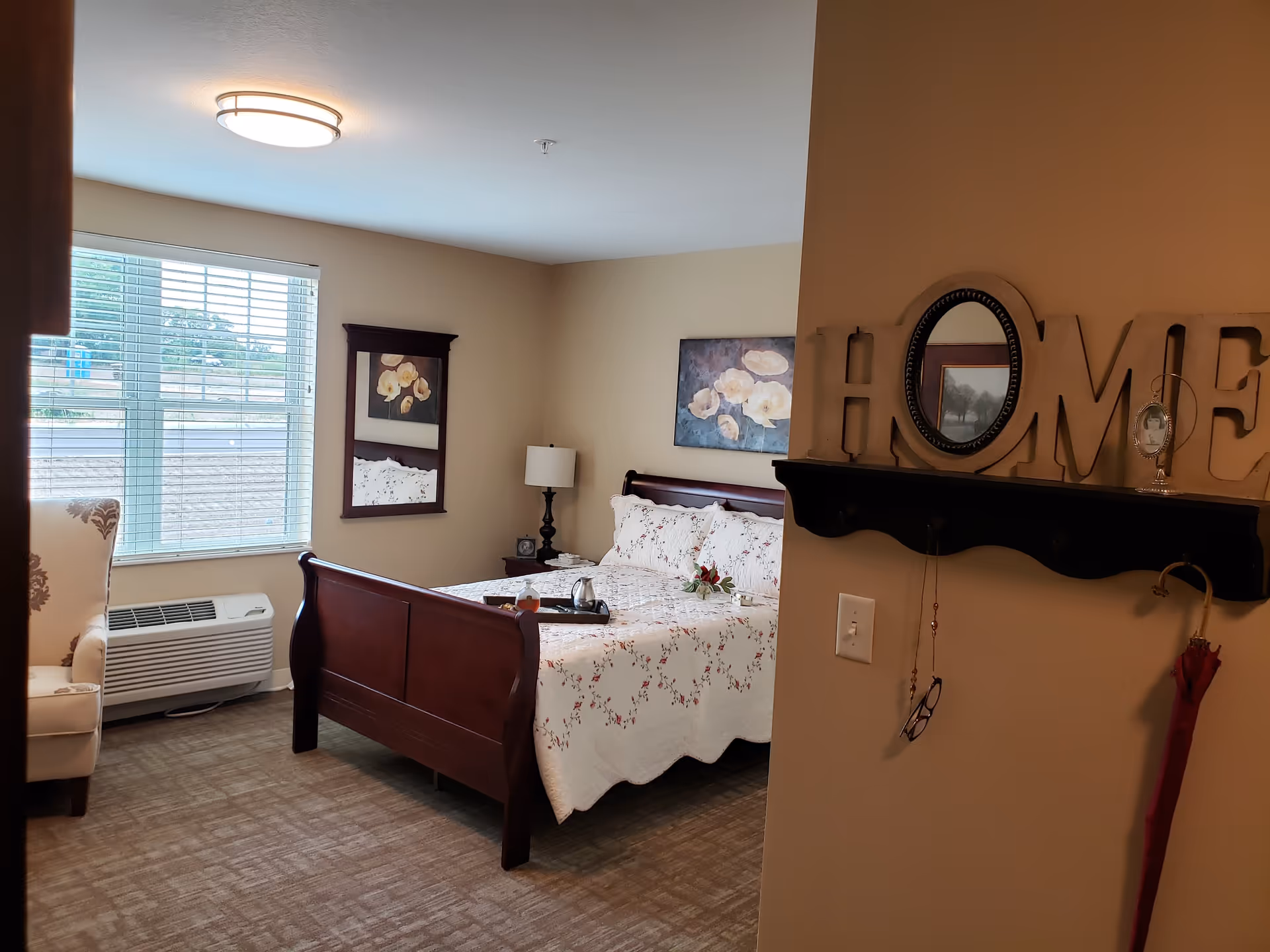 A cozy bedroom with a wooden bed frame and floral bedspread. There is a window with blinds letting in natural light, a cushioned armchair with a floral pattern, a nightstand with a lamp, and a wall-mounted mirror. On the right side, a wall shelf holds decorative letters spelling 'HOME' with a small oval mirror in the letter O, a framed photo, and hanging items including glasses and an umbrella.