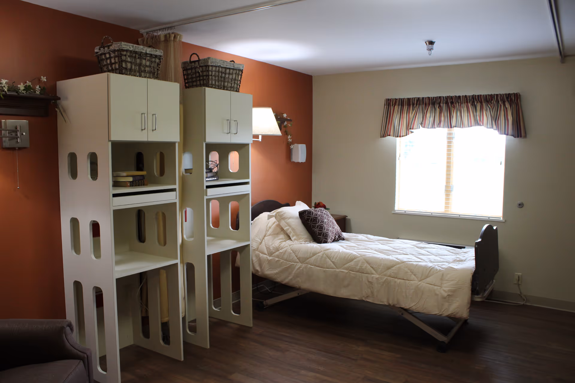 A senior living facility bedroom with a single bed covered in a white quilt and a decorative pillow. The room has two tall white shelving units with baskets on top, a window with striped valance curtains, and a brown accent wall with a wall lamp. The floor is wood, and there is a brown armchair partially visible in the corner.