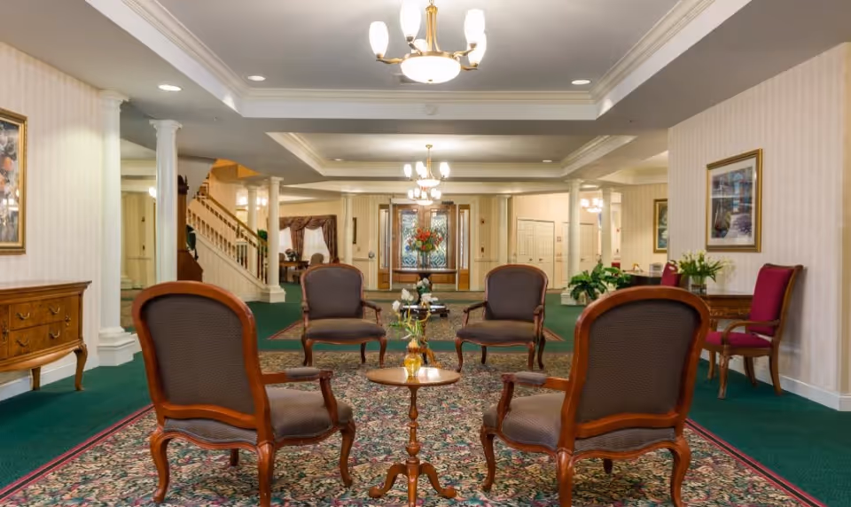 A spacious and elegant common area with vintage-style wooden chairs arranged around small tables on a patterned carpet. The room features green carpeting, cream-colored walls with framed artwork, white columns, and multiple chandeliers hanging from a tray ceiling. In the background, there is a staircase and a set of double doors with decorative glass panels.