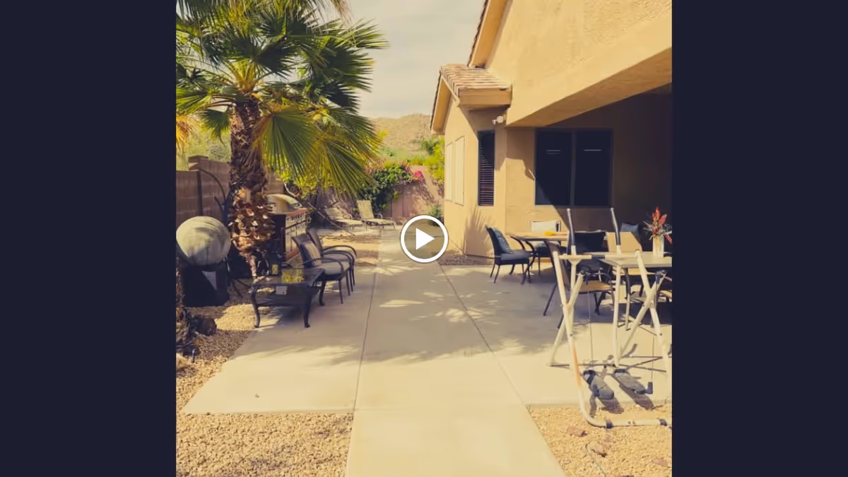 Sunlit outdoor patio and walkway beside a beige assisted-living building with chairs, tables, palm trees, and a mobility walker.