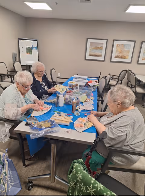 Three elderly women sitting around a table in a room, engaging in a craft activity involving decorative hand fans and various craft supplies spread out on a blue tablecloth. The room has beige walls with framed artwork and several empty chairs in the background.