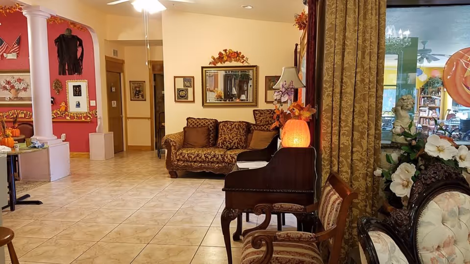 Interior view of a common area in East Lake Manor Assisted Living Facility featuring a patterned sofa, a wooden piano with a glowing lamp on top, ornate chairs, floral decorations, and a large mirror on the wall. The room has tiled floors, warm lighting, and autumn-themed decorations.
