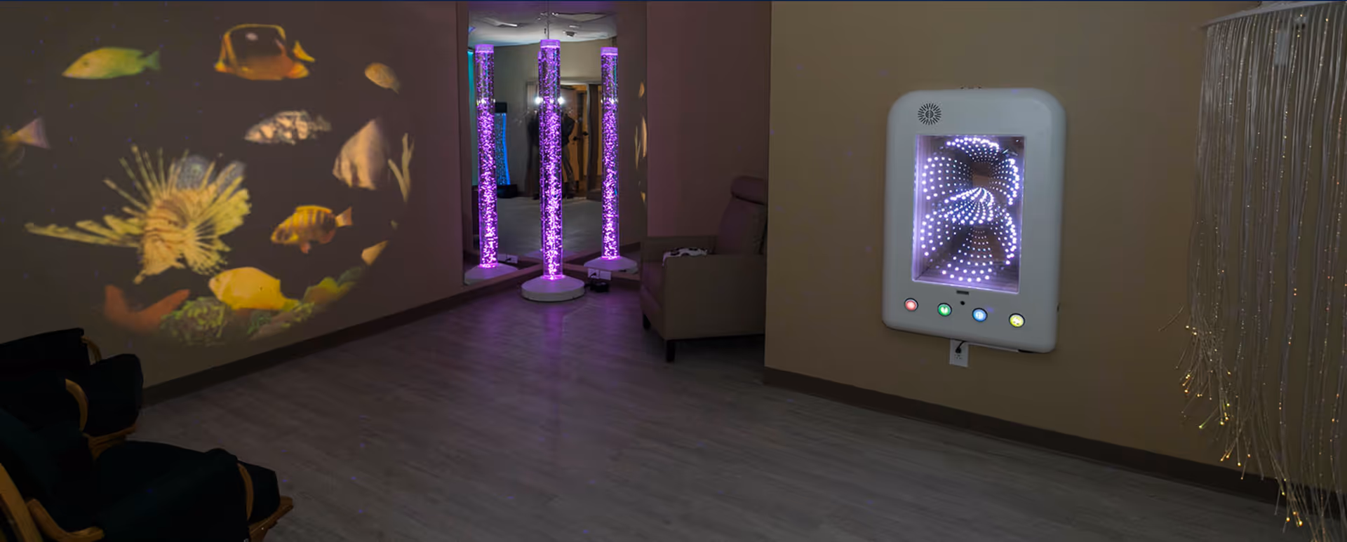 A sensory room with dim lighting featuring a wall projection of colorful fish, three vertical bubble tubes illuminated with purple light in front of a mirror, a wall-mounted infinity mirror light, fiber optic strands hanging on the right, and a comfortable chair in the corner.