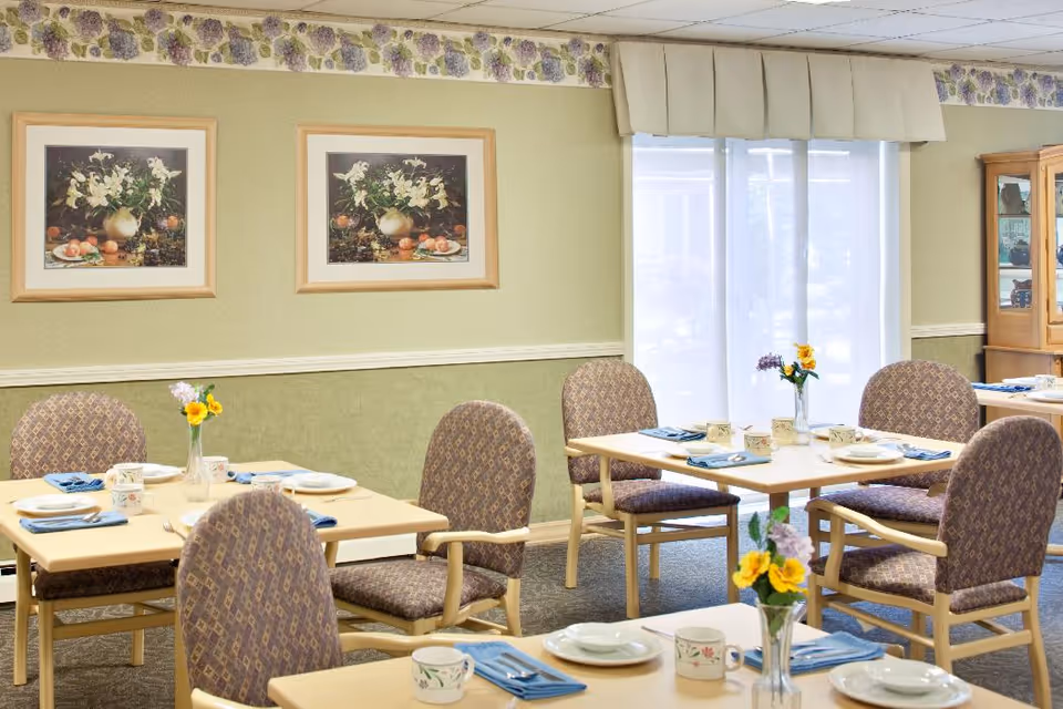 A dining room in a senior living facility with several tables set for a meal. Each table has plates, cups, utensils, and napkins neatly arranged. There are patterned upholstered chairs around the tables. The room has light green walls with a floral border near the ceiling, two framed floral paintings on the wall, and a large window with sheer white curtains letting in natural light. A wooden cabinet with glass doors is visible in the background.