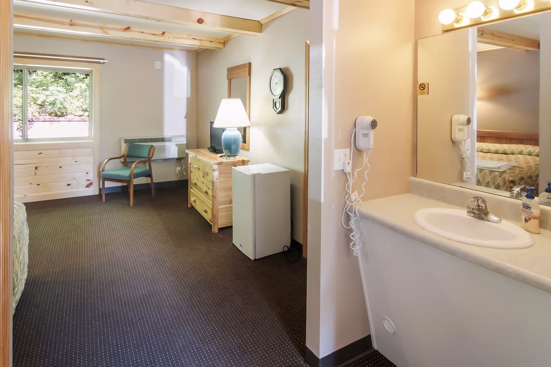 Small assisted-living bedroom with a bed, dresser, lamp and chair, plus a sink vanity and mirror in the foreground.