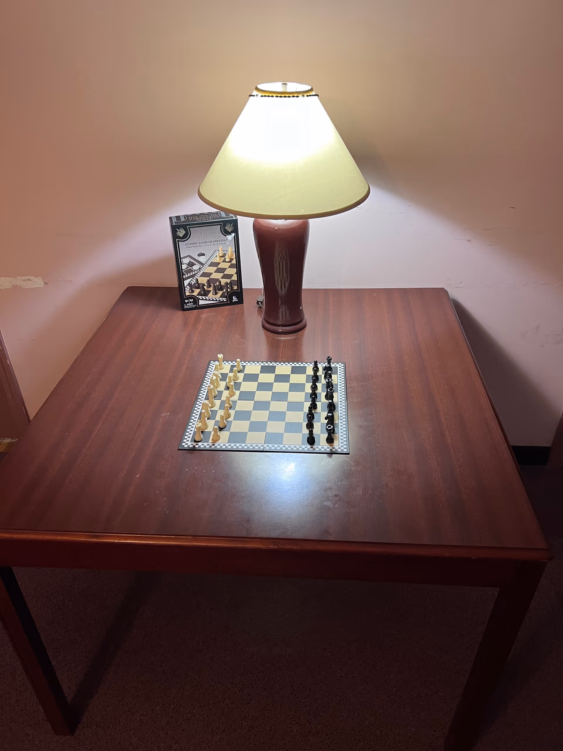 A wooden table with a chessboard set up for a game, illuminated by a table lamp with a beige lampshade. A chess instruction booklet is propped up behind the lamp against a plain wall.