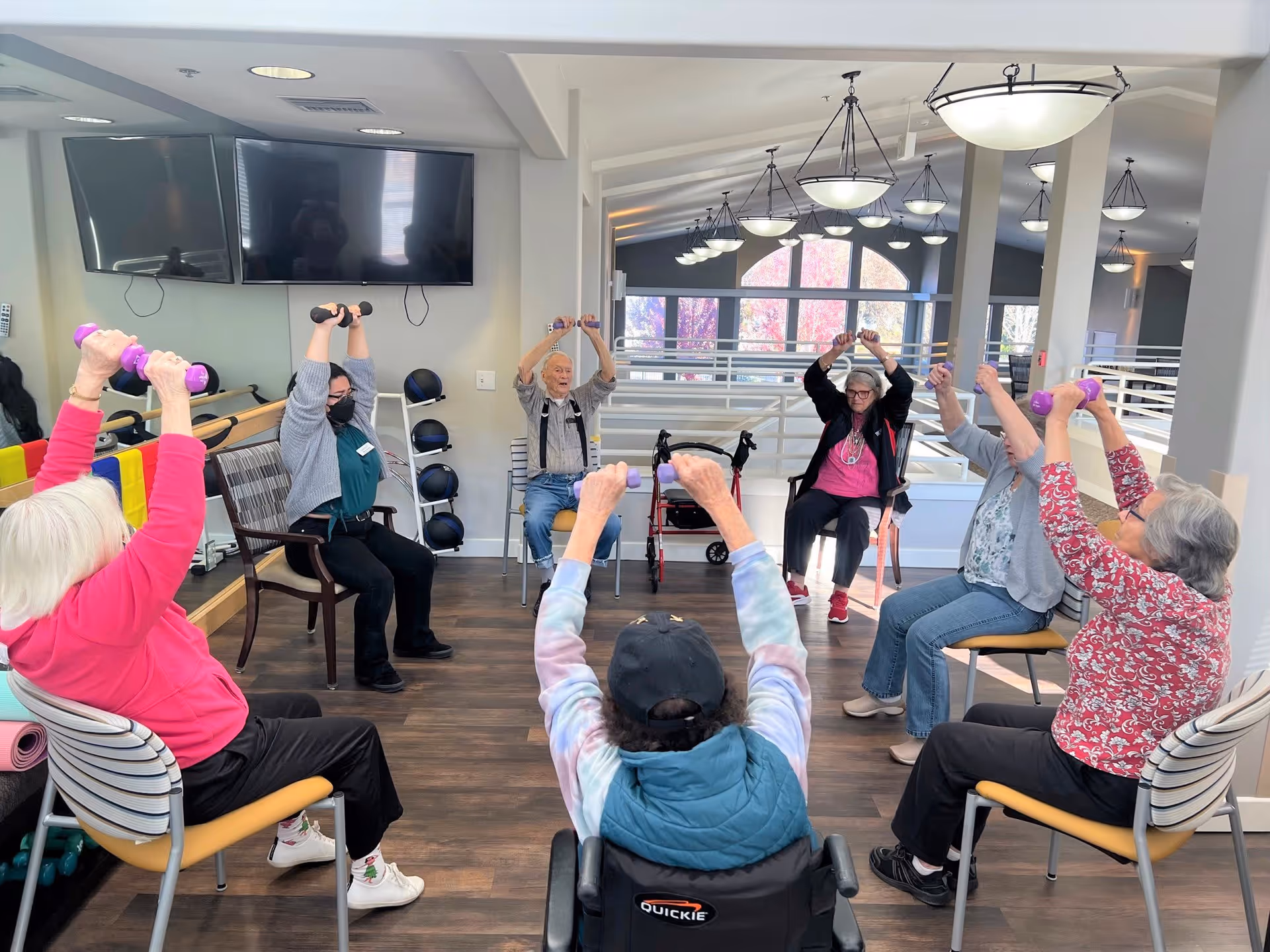 A group of elderly individuals seated in a circle in a bright, spacious room participating in a seated exercise class, lifting small dumbbells overhead. A staff member wearing a mask is also participating. The room has large windows, modern lighting, and two large TVs mounted on the wall.