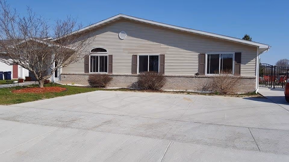 Single-story beige building front with three windows, shrubs, a leafless tree and a wide concrete driveway under a clear blue sky.