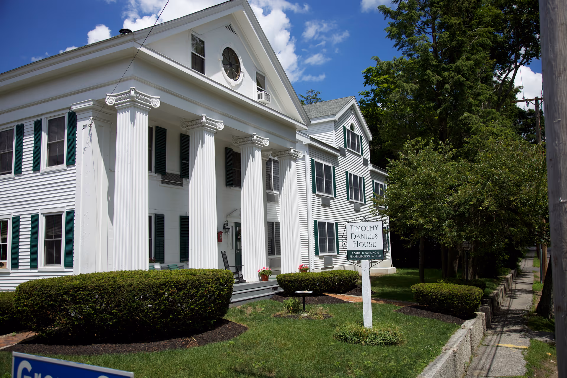 White colonial-style building with large columns and a yard sign that reads "Timothy Daniels House" under a blue sky.