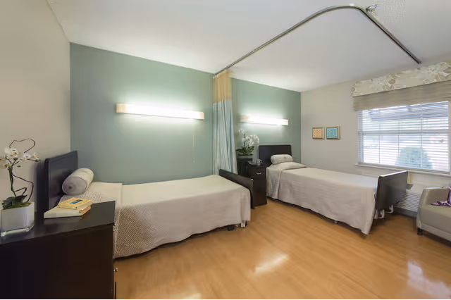 A bright and clean room with two single beds separated by a nightstand. Each bed has a light beige bedspread and a cylindrical pillow. The walls are painted light green and beige, with modern horizontal wall lights above each bed. There is a window with blinds and a floral valance on the right side, and a beige armchair next to it. The floor is light wood, and there is a ceiling-mounted curtain track for privacy between the beds. A small plant and a book are on the nightstand.