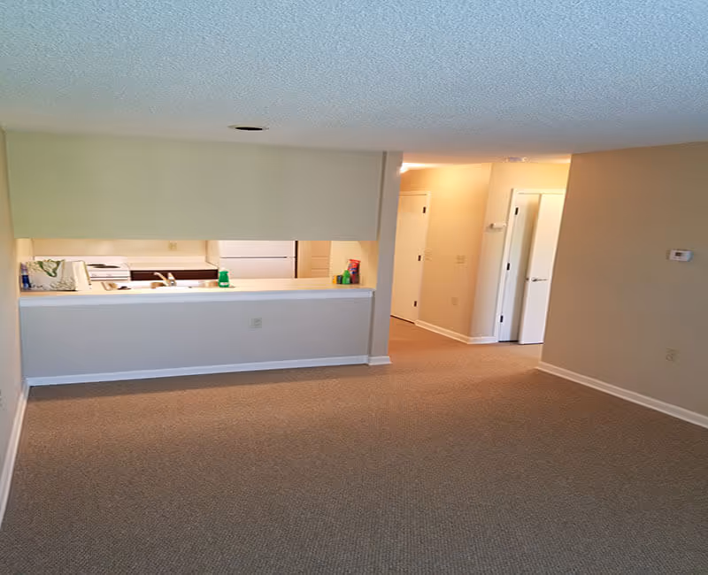 Interior view of an empty apartment with beige carpet and walls. The image shows a small kitchen area with a white refrigerator, stove, and sink, partially visible through a pass-through window. There is a hallway leading to closed doors on the right side.