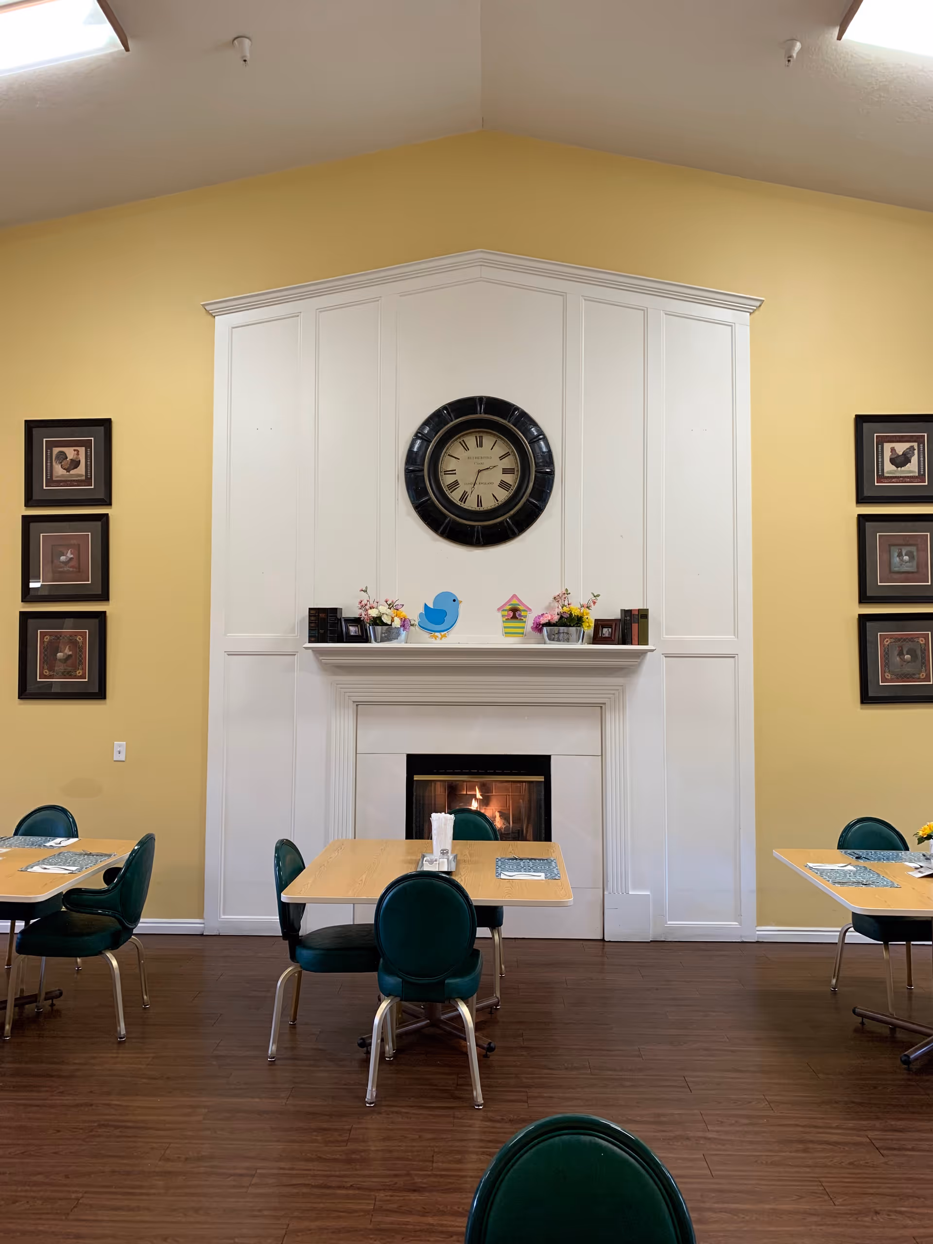 Dining area with wooden tables and green chairs arranged around a white fireplace with a lit fire. Above the fireplace is a large round clock and decorative items including flowers and small birdhouse figurines. The walls are painted yellow and decorated with framed pictures of roosters.