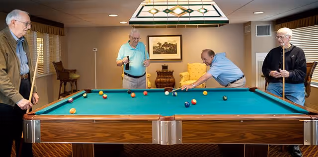 Four elderly men playing a game of pool in a well-lit room with beige walls, a green pool table, and a hanging light fixture above the table. The room has windows with blinds and a framed picture on the wall.