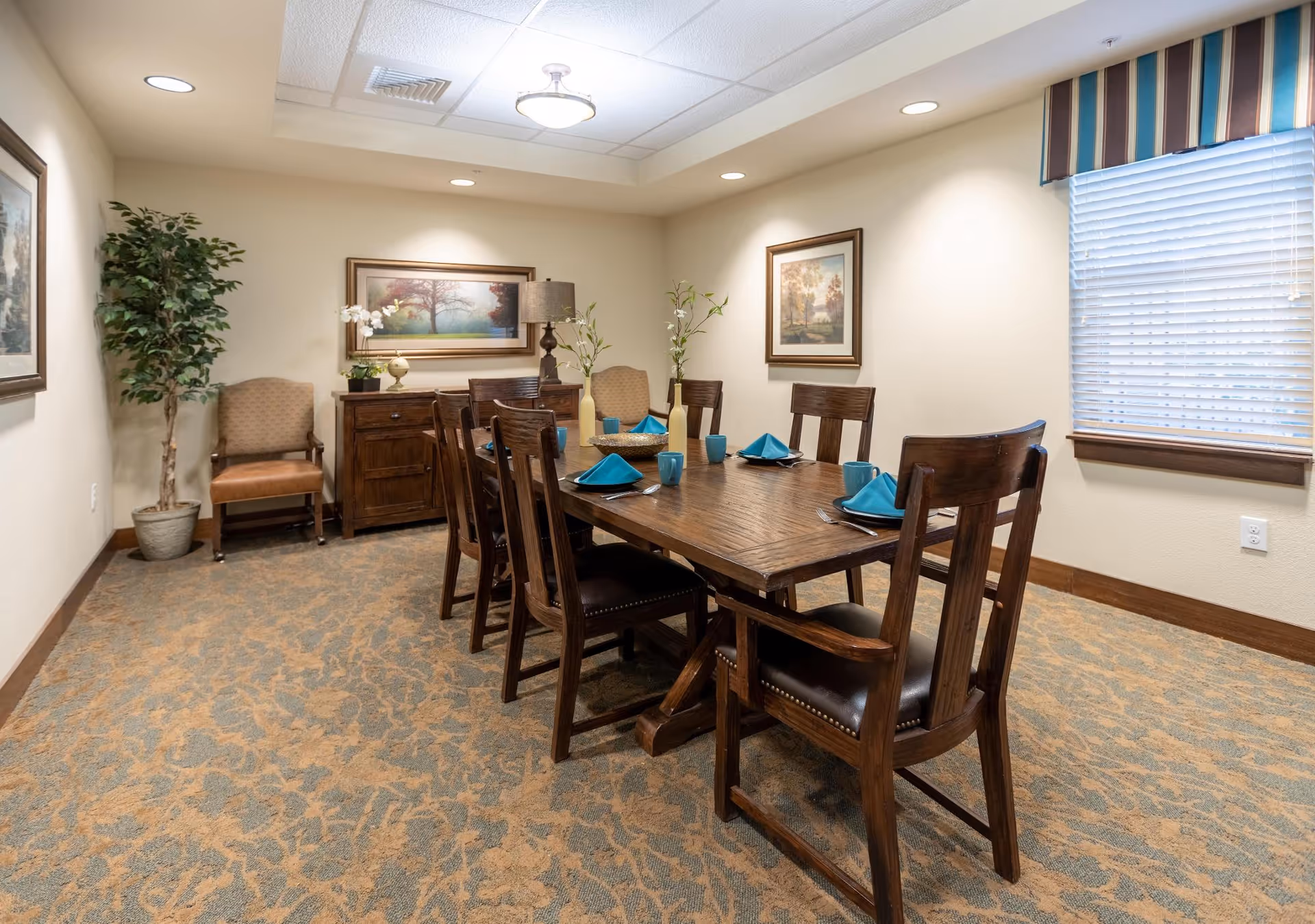 A dining room with a wooden table set for six with blue napkins and matching cups. The room has beige walls, carpeted floor, two framed landscape paintings, a window with blinds and a striped valance, a wooden sideboard with a lamp and plants, and two upholstered chairs in the corner.