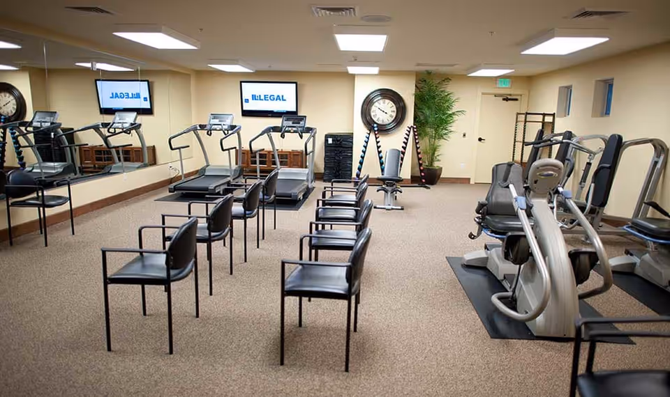 A fitness room with exercise equipment including treadmills, stationary bikes, and weight machines. Several black chairs are arranged in rows in the center of the room. A large mirror covers one wall, reflecting the equipment and chairs. A clock and a potted plant are visible on the far wall beneath ceiling lights.