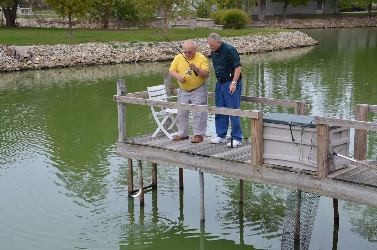 Two elderly men fishing off a wooden dock on a greenish pond. One man in a yellow shirt is holding a fishing rod with a fish caught on the line, while the other man in a dark green shirt watches. There is a white chair and a storage box on the dock, with trees and rocks visible on the far shore.
