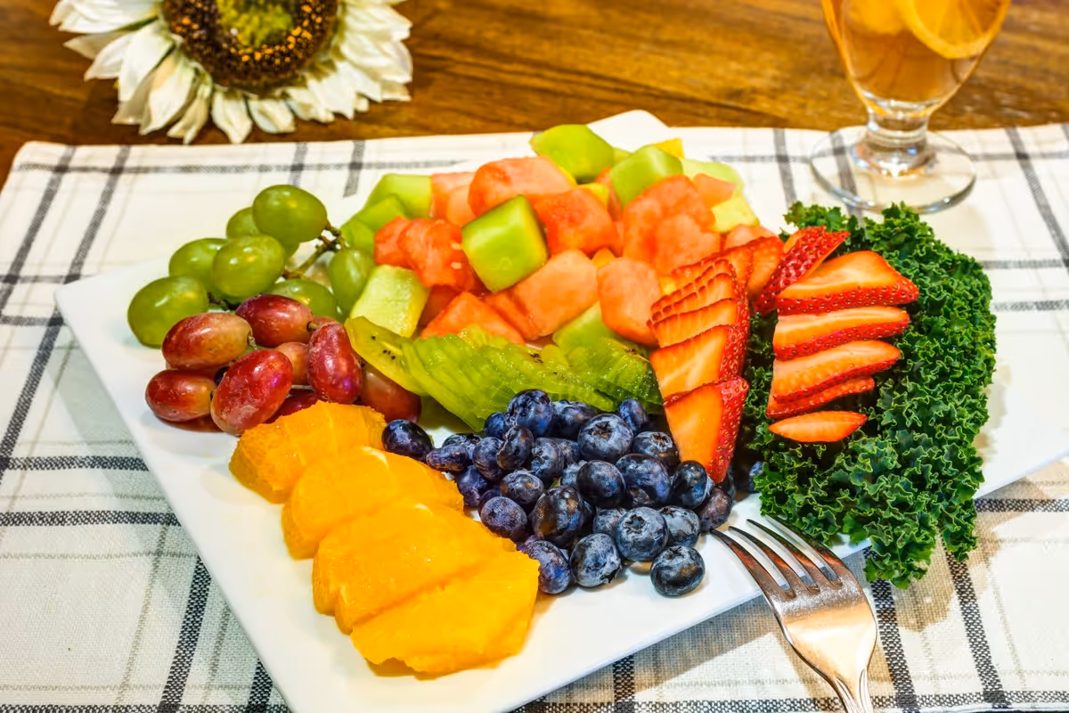 A white square plate filled with a colorful assortment of fresh fruits including green grapes, red grapes, orange slices, blueberries, kiwi slices, watermelon chunks, and sliced strawberries, garnished with curly kale. A fork rests on the plate and a glass of iced tea is visible in the background on a checkered tablecloth.