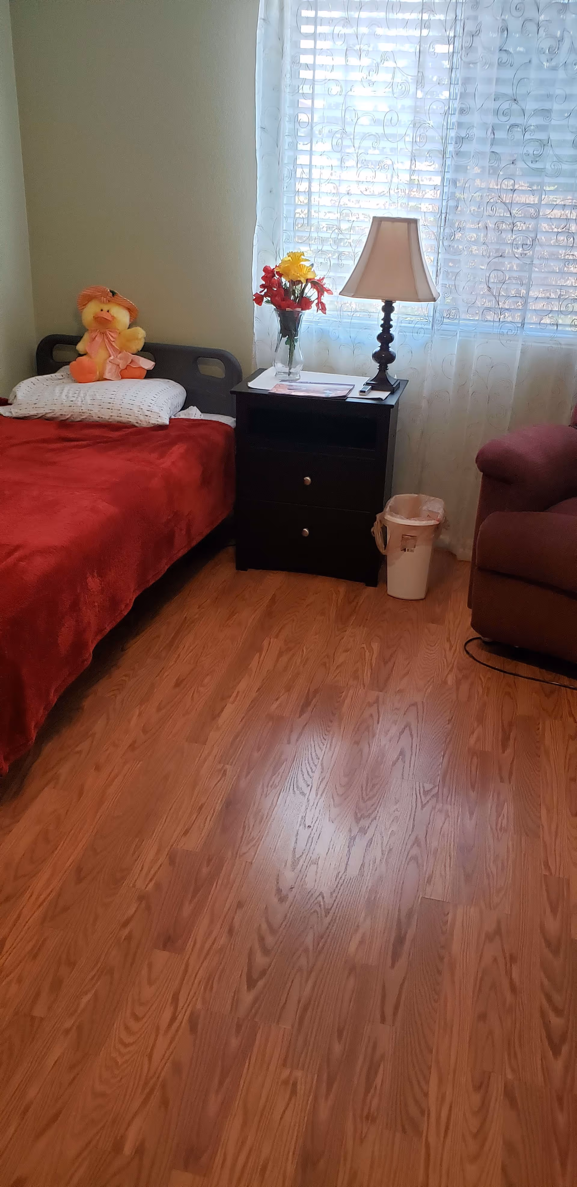 A cozy bedroom with a single bed covered in a red blanket and a white pillow with a stuffed duck toy on it. Next to the bed is a black nightstand with two drawers, a vase with yellow and red flowers, some papers, and a table lamp. A window with sheer white curtains is behind the nightstand. There is a maroon armchair partially visible on the right side and a small trash bin on the floor beside the nightstand. The floor is wooden.