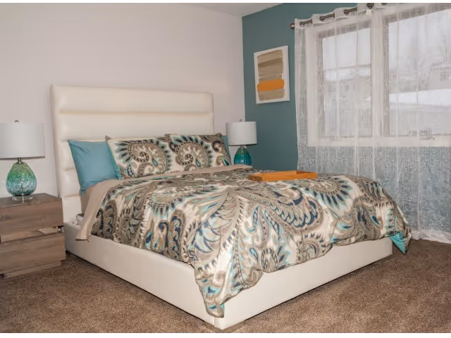 A bedroom with a large white upholstered bed featuring a patterned comforter in shades of teal, beige, and brown. There are two matching pillows and two teal pillows. On each side of the bed is a wooden nightstand with a teal glass lamp. A window with sheer white curtains is on the right wall, and a framed abstract artwork hangs on the teal accent wall behind the bed.