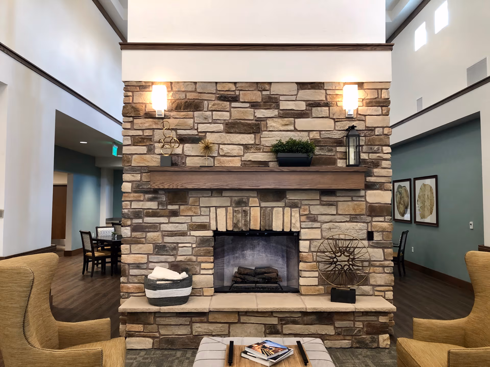 A cozy common area with a large stone fireplace in the center, flanked by two yellow armchairs. The fireplace mantel holds decorative items including a small plant and a lantern. Behind the fireplace, there are tables and chairs, and framed artwork on the walls. The room has high ceilings with windows near the top, allowing natural light to enter.
