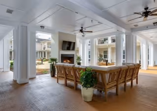 Covered outdoor communal seating area with a long wooden table and chairs facing a fireplace and TV, ceiling fans overhead, and views into a courtyard and adjoining building.