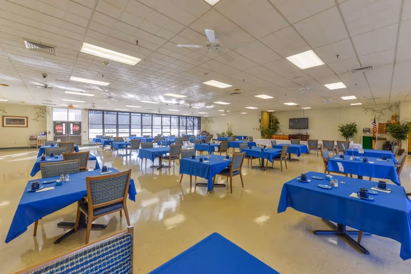 A spacious dining room with multiple tables covered in blue tablecloths, each set with blue cups, saucers, and silverware. The room has large windows letting in natural light, ceiling fans, and a television mounted on the far wall. There are plants and an American flag near the back corner of the room.