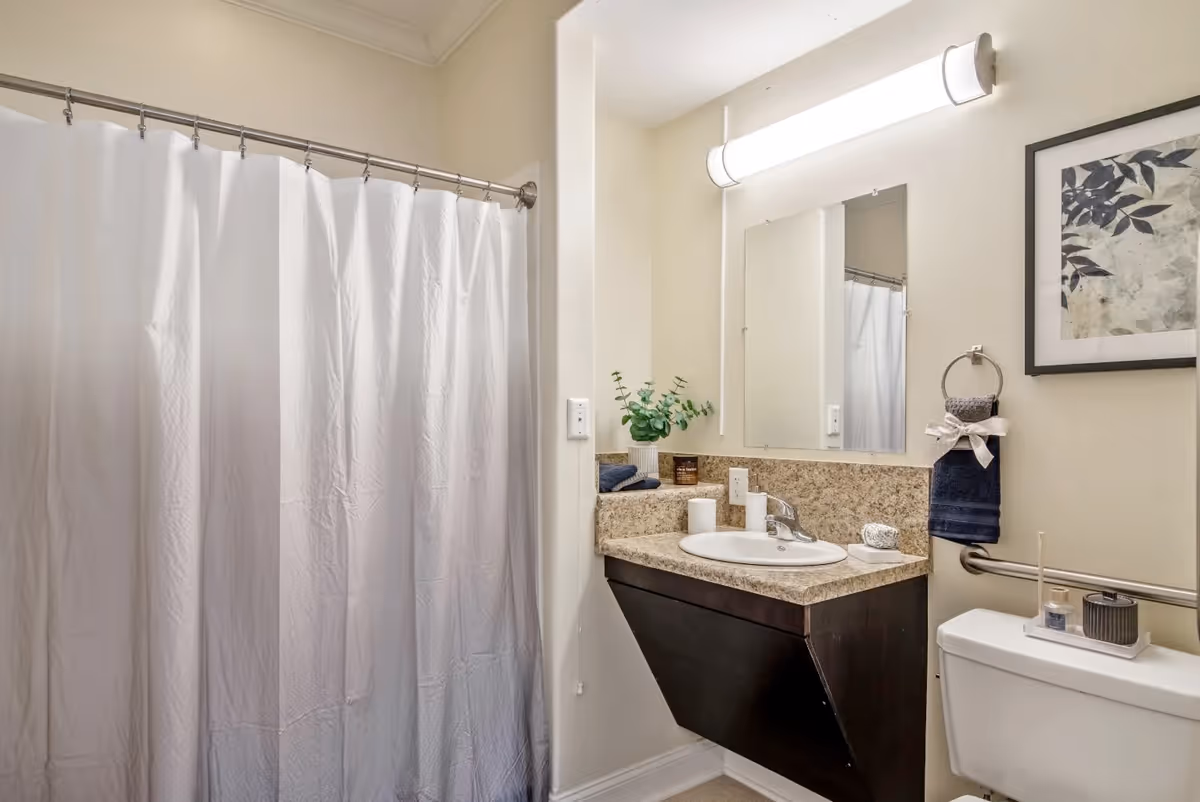 A clean bathroom featuring a white shower curtain on a curved rod, a countertop with a sink, a mirror above the sink, a wall-mounted light fixture, a toilet with a tray holding decorative items, a towel ring with a dark blue towel tied with a ribbon, and a framed artwork on the wall.