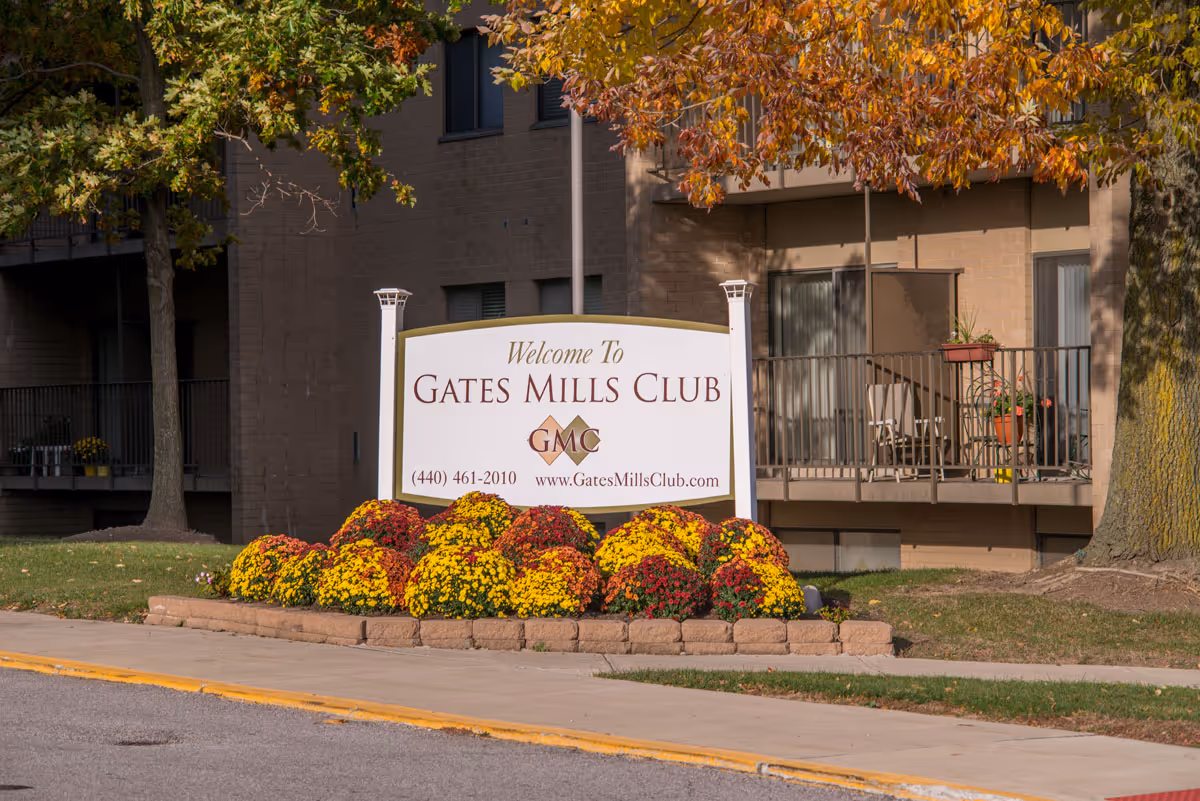 Outdoor view of the entrance sign for Gates Mills Club Senior Living, surrounded by colorful flowers and trees with autumn foliage, with a building and balconies in the background.