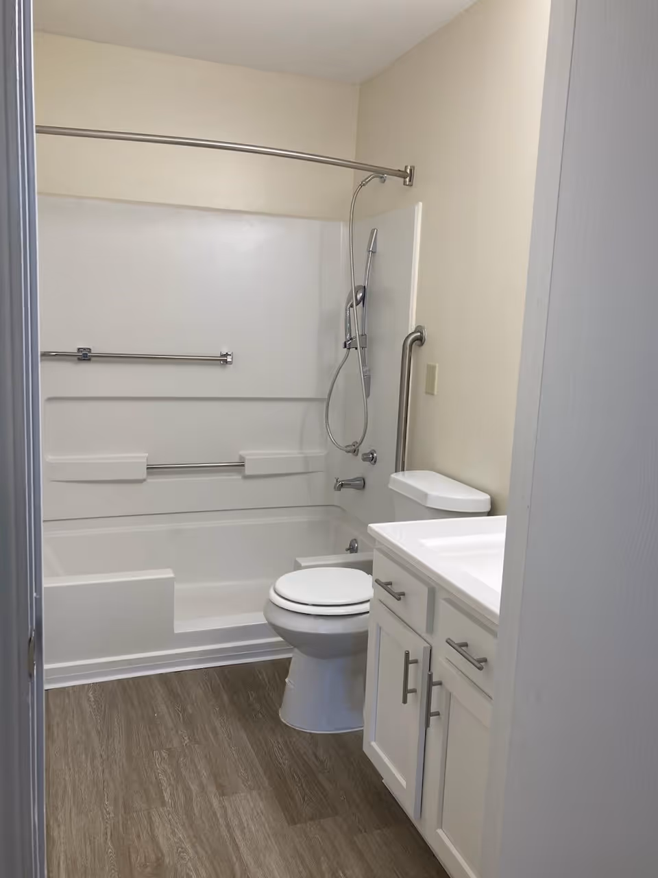 A clean and modern bathroom featuring a white bathtub with a showerhead and grab bars, a white toilet, and a white vanity with a sink and storage cabinets. The floor has wood-like vinyl flooring and the walls are painted a light beige color.