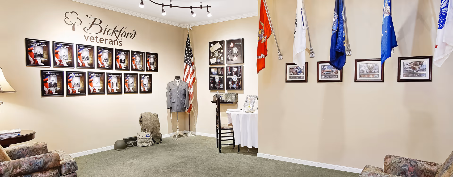 Interior common room displaying a 'Bickford veterans' wall with framed plaques, flags, and seating.
