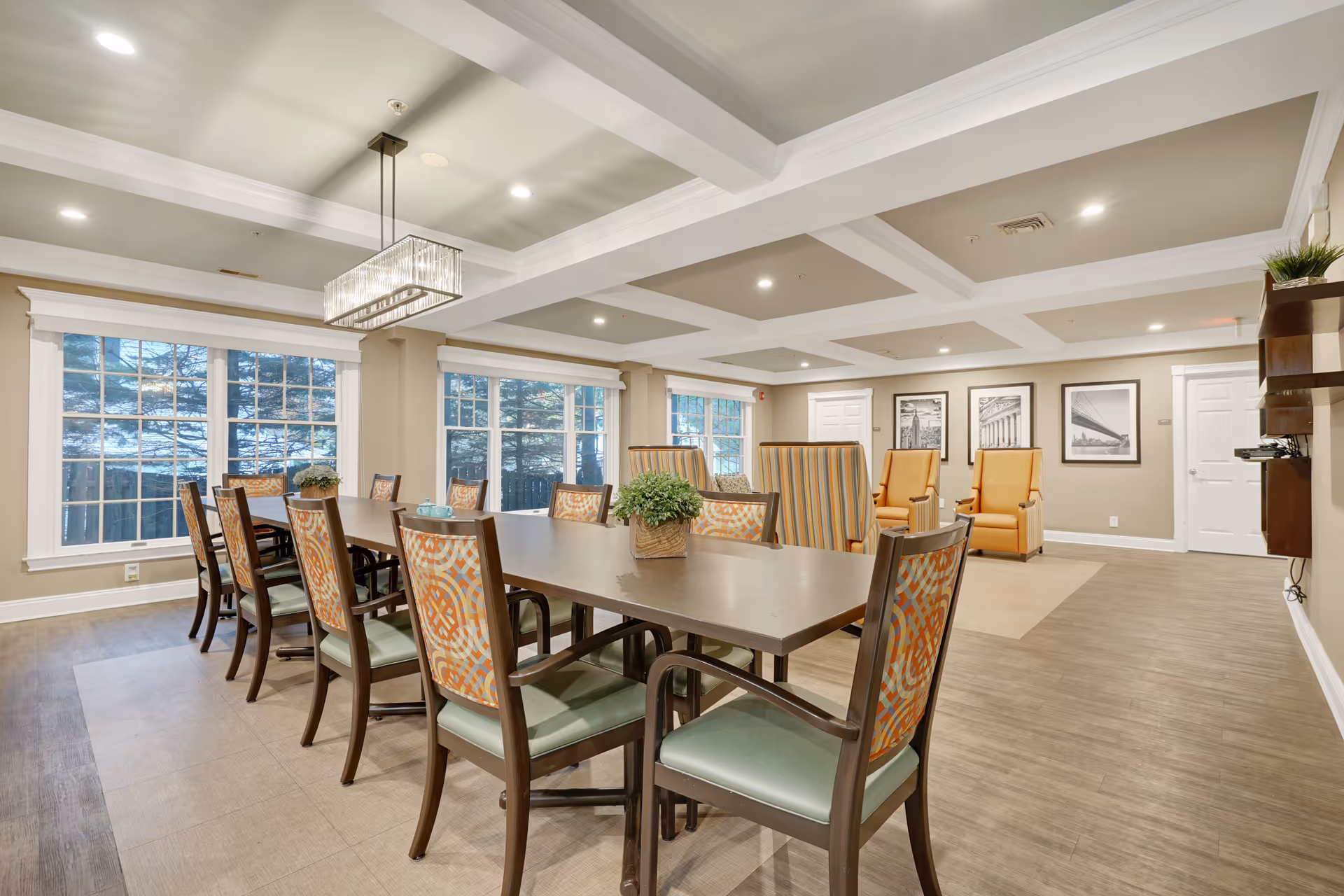 A spacious, well-lit common area in a senior living facility featuring a long dining table with patterned chairs, large windows letting in natural light, a modern rectangular chandelier, and a seating area with comfortable armchairs and framed black-and-white photographs on the wall.