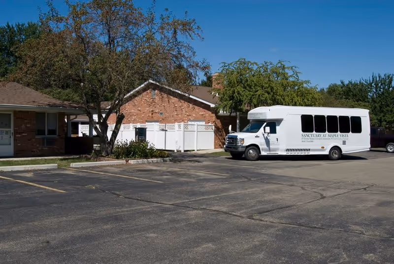 A white shuttle bus parked in front of a single-story brick building with a tree and a white fence. The bus has the text 'SANCTUARY AT MAPLE VISTA' on its side. The scene is set in a parking lot under a clear blue sky.