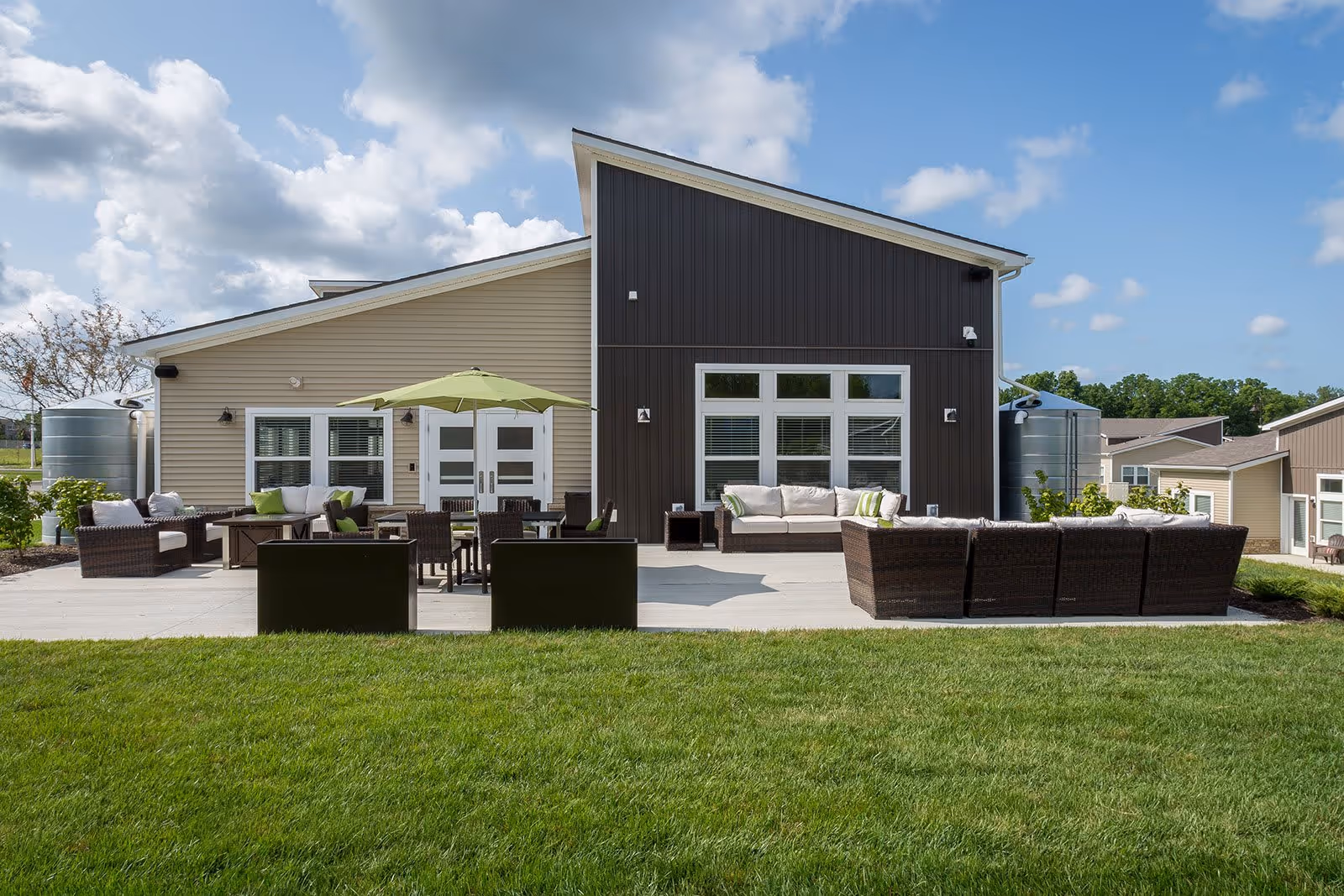 Modern single-story building facade with a paved patio, outdoor seating, umbrella, and a manicured lawn under a partly cloudy sky.