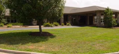 A single-story senior living facility entrance with brick exterior, green lawn, and trees.