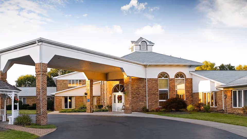 Exterior view of a senior living facility with a covered entrance supported by brick columns, large windows, and a well-maintained lawn under a partly cloudy sky.