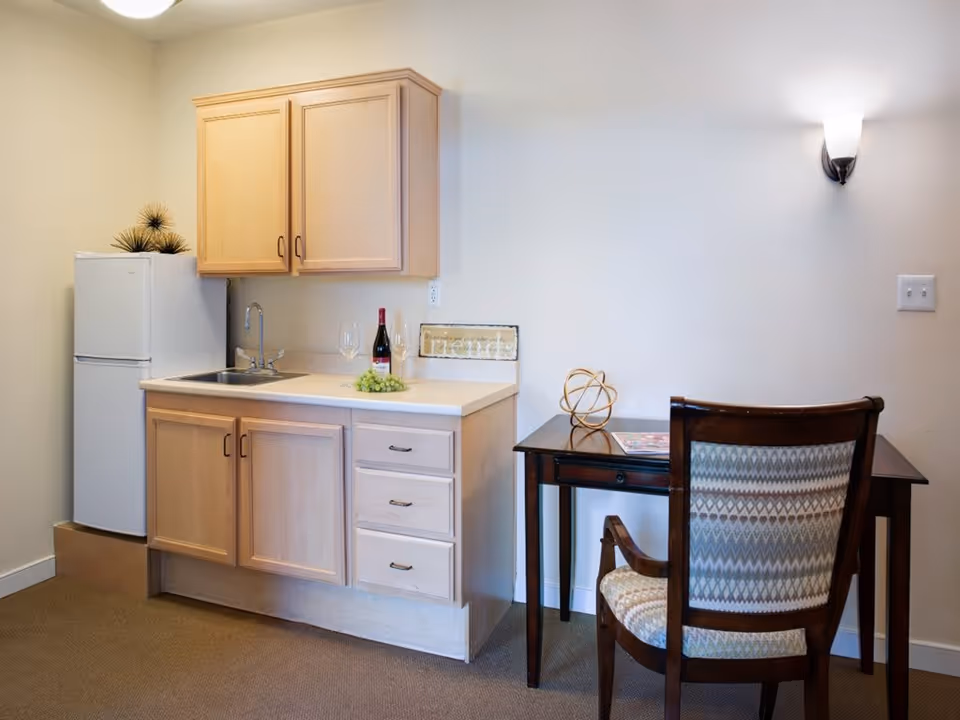 A small kitchenette area with light wood cabinets, a white refrigerator, a sink, and a countertop with a bottle of wine, two wine glasses, and a small bunch of grapes. Next to the kitchenette is a dark wooden desk with a decorative object and a magazine on it, accompanied by a wooden chair with patterned upholstery. The walls are light-colored with a wall-mounted light fixture above the desk.