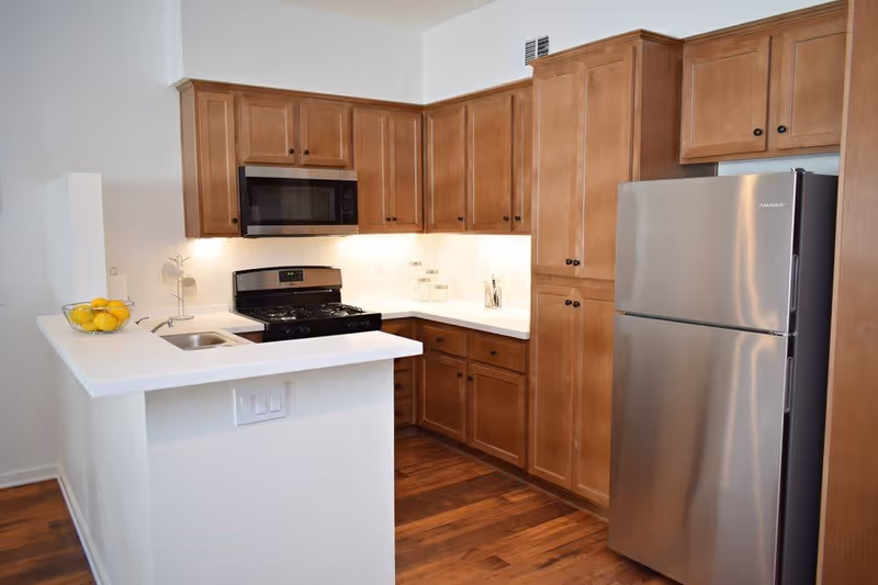 Modern kitchen with wooden cabinets, stainless steel refrigerator, microwave, and stove. The kitchen has a white countertop with a small sink and a bowl of lemons on the counter. The floor is wooden and the walls are white.