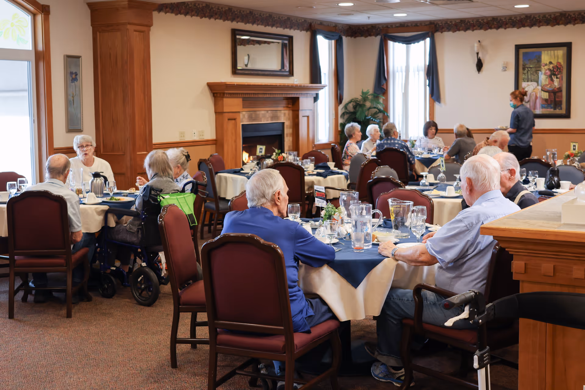 A dining room in The Winslow Independent Living facility with elderly residents seated at round tables covered with blue and white tablecloths. The room features a fireplace, framed artwork, and large windows with blue curtains. A staff member wearing a mask is attending to the residents.