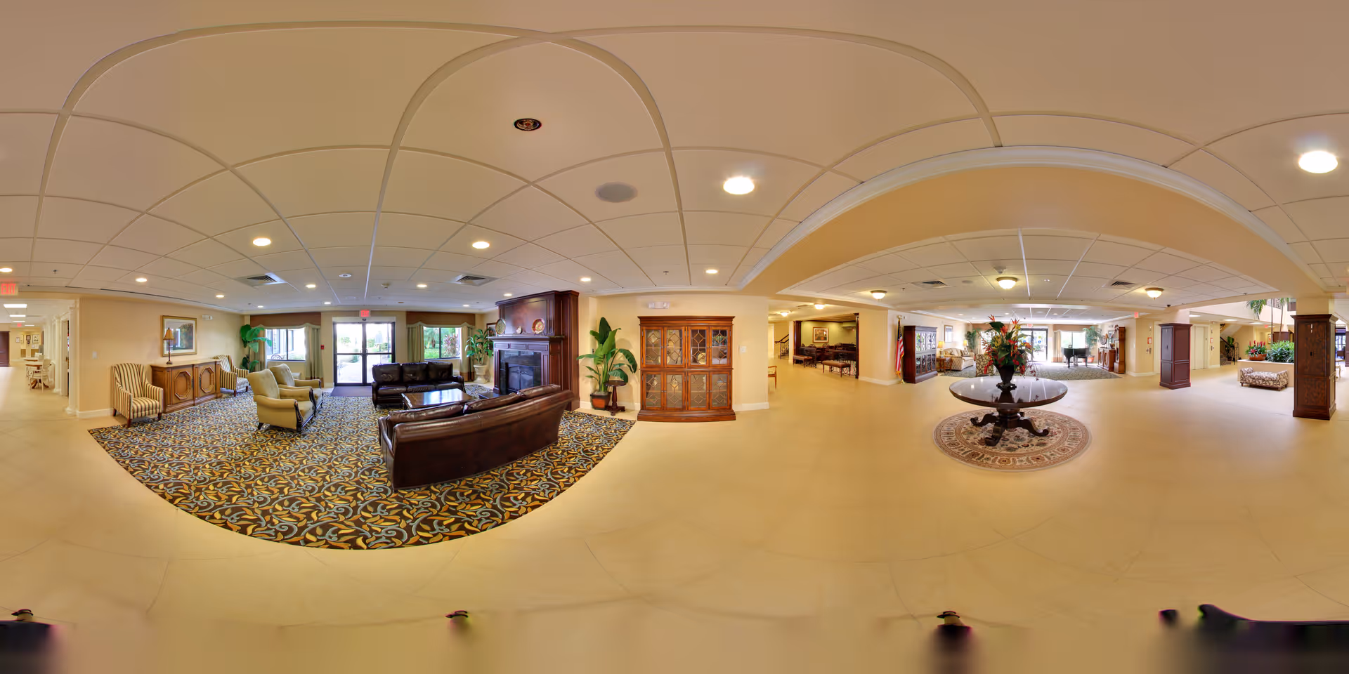 A spacious and well-lit senior living facility common area with a seating arrangement including leather sofas and armchairs around a fireplace. The room features patterned carpet and tiled flooring, decorative plants, wooden furniture, and a round table with a floral centerpiece on a circular rug. Large windows and glass doors allow natural light to enter, and the ceiling has recessed lighting.