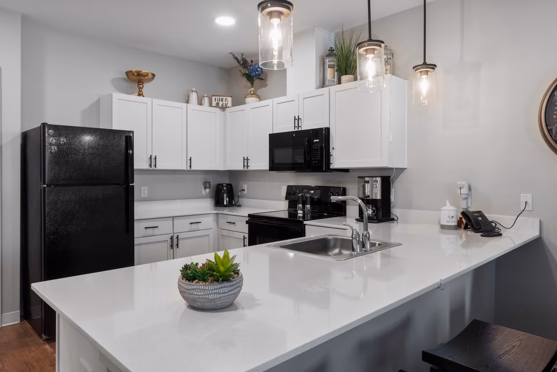 Modern kitchen with white cabinets, black refrigerator, black microwave, black stove, and a white countertop island with a small potted succulent plant. Three pendant lights hang above the island, and there is a sink with a faucet on the island. Various kitchen appliances and decorative items are on the countertops and cabinets.