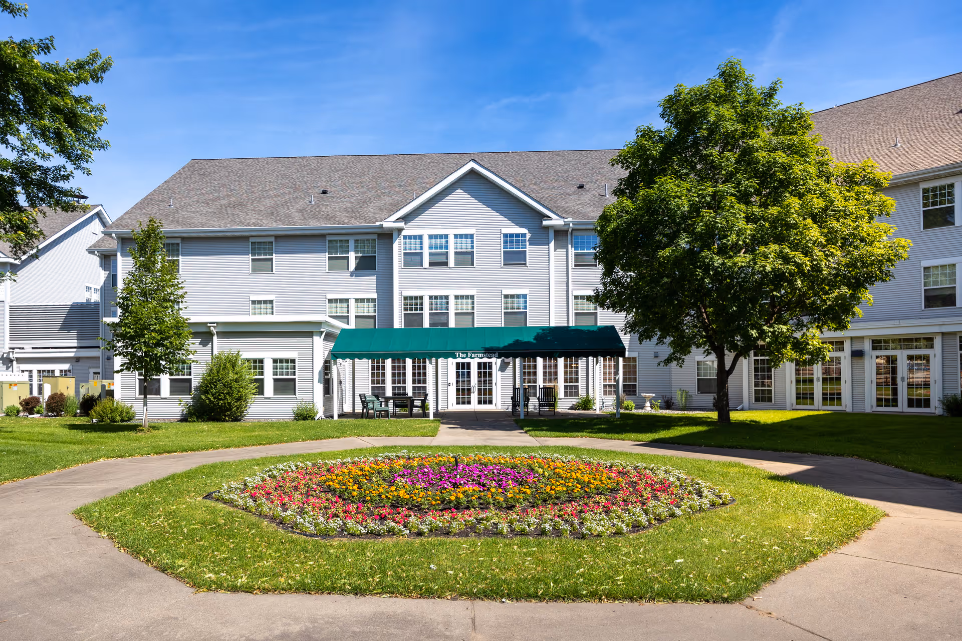 Exterior view of The Farmstead senior living facility showing a three-story building with multiple windows, a green awning over the entrance, a circular flower bed with colorful flowers in the foreground, and trees on a sunny day with a clear blue sky.
